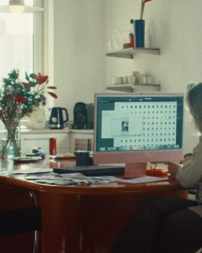 Persoon werkt aan een roze iMac aan een rode tafel in een lichte keuken, beeld uit een campagne
